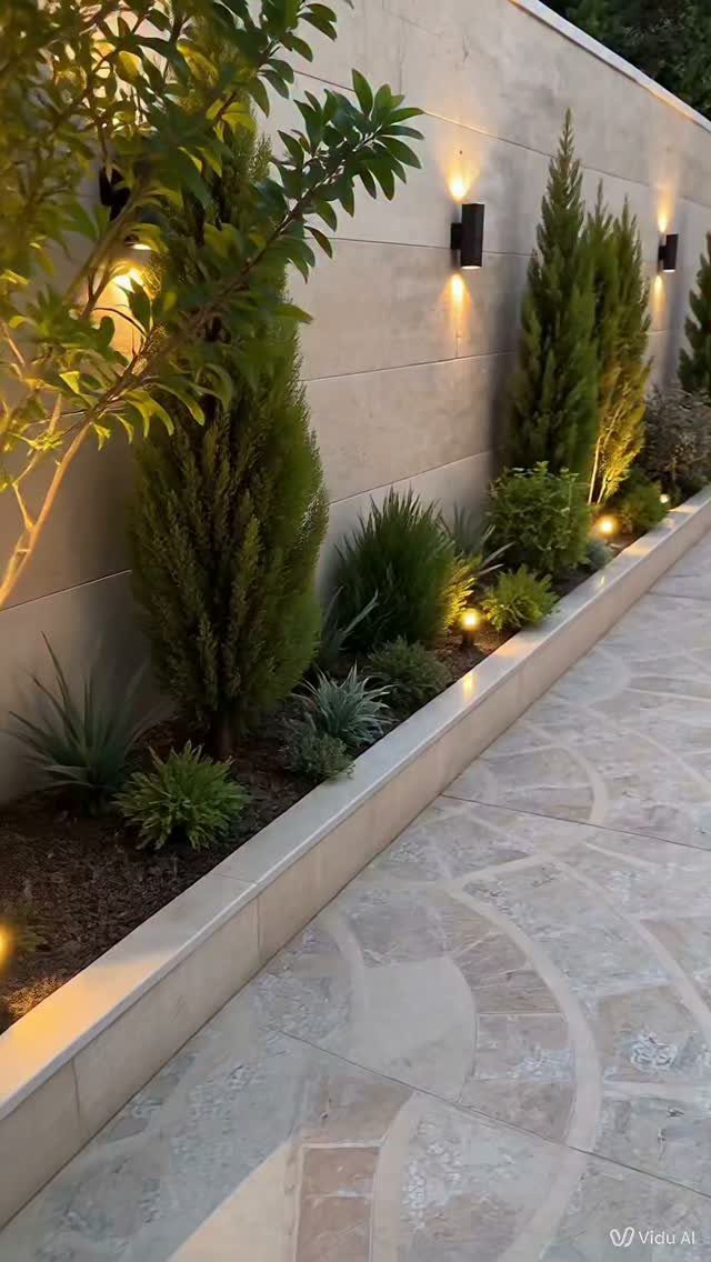 19 Small Garden Design Ideas with Built-In Planning Tips 17 Tall evergreen shrubs and green plants illuminated by warm wall-mounted lights along a stone path.