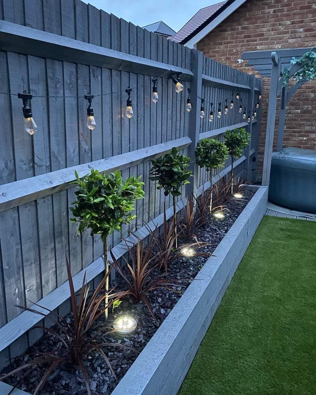 12 Tiny Garden Ideas for Patios, Balconies and Small Yards 4 String lights hanging along a grey wooden fence above a narrow garden bed with small shrubs.