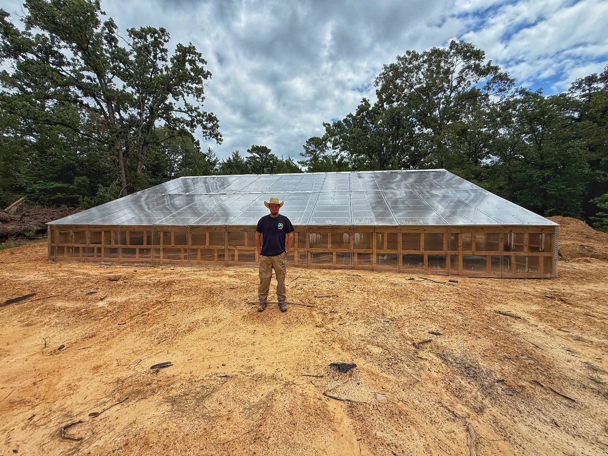 Under Ground Greenhouse: 5 Ways to Grow Below the Surface 2 A man standing in front of a large, low-profile greenhouse under construction in a clearing.